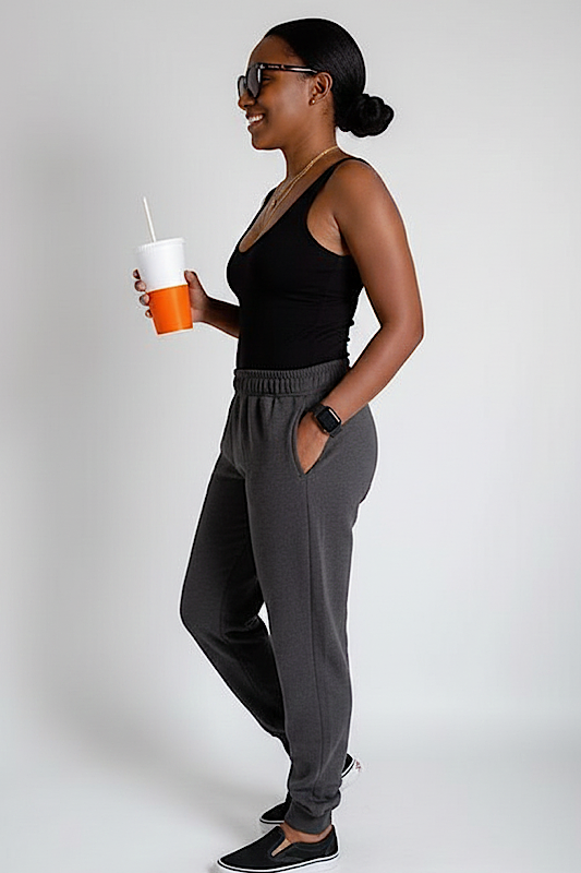 Woman in black top and gray pants holding a drink against a plain background